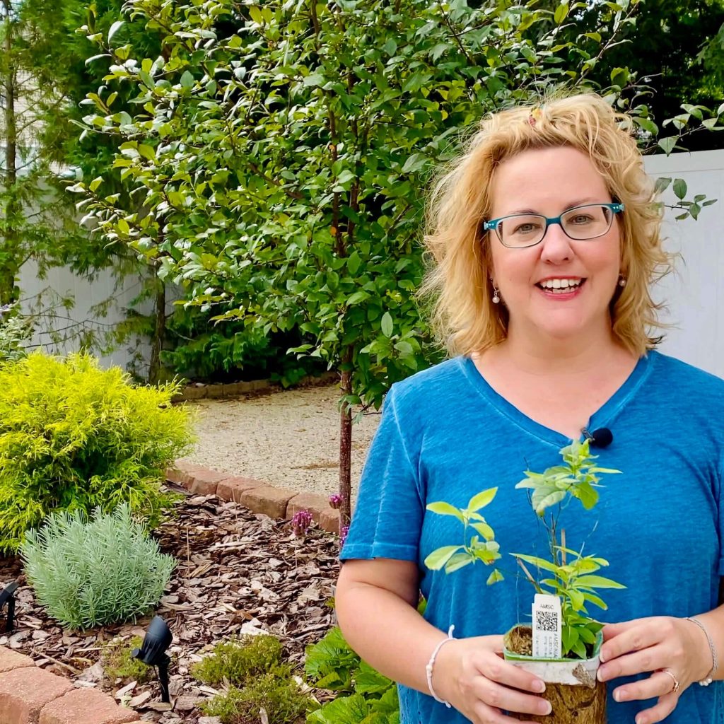 Laura from Garden Sanity smiling and holding a seedling plant in front of a garden bed